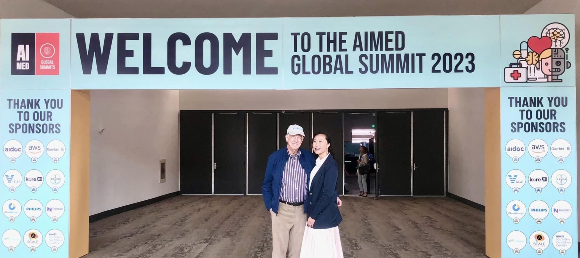 Sherri Douville and Art Douville standing together beneath the AIMed Global Summit 2023 welcome banner at the conference entrance.